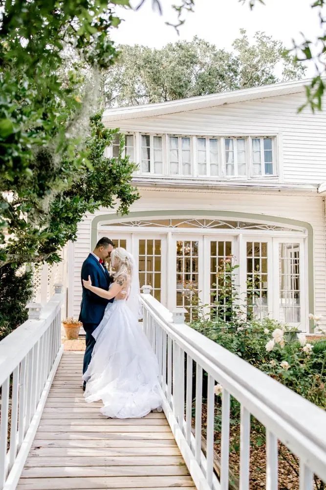 Bride and groom embracing on a white wooden bridge in front of a large house with multiple windows.