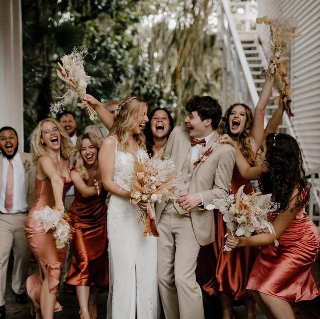 Group of people celebrating at a wedding, with the bride in a white dress and bridesmaids in peach-colored dresses, outdoors.