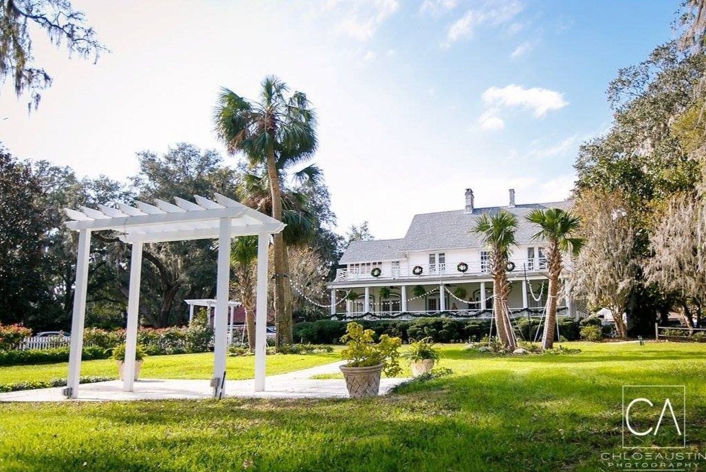Historic white house with a large front porch, surrounded by palm trees and greenery, with a white pergola in the foreground under a blue sky.