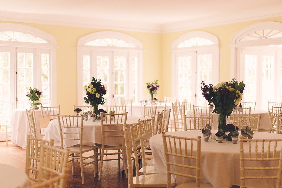 Elegant, well-lit room with round tables, white tablecloths, and flower centerpieces. Light tan chairs are arranged around each table, and tall windows allow natural light to fill the space.