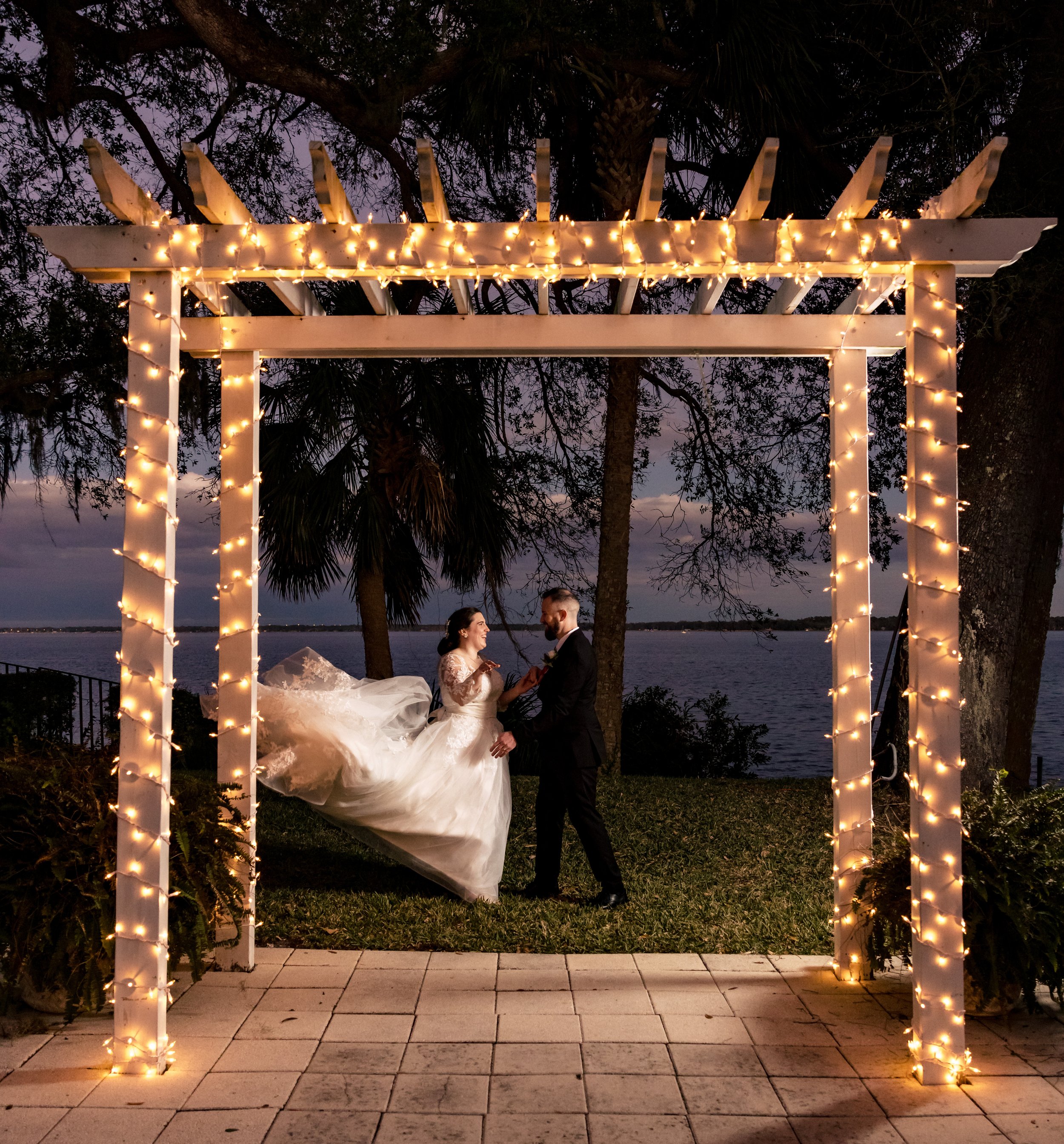 A bride and groom posing under a pergola decorated with string lights, outdoors near a body of water.