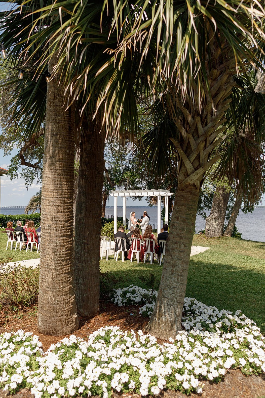 Outdoor wedding ceremony under a gazebo by the water, with guests seated and palm trees and flowers in the foreground.