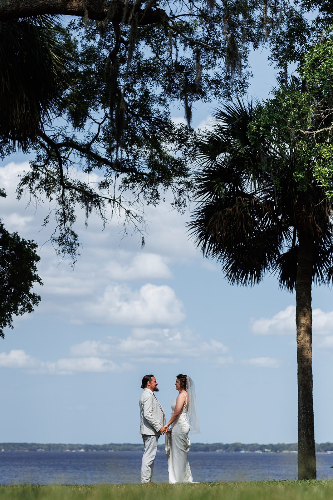 Bride and groom holding hands outdoors by a lake, surrounded by trees, clear sky.