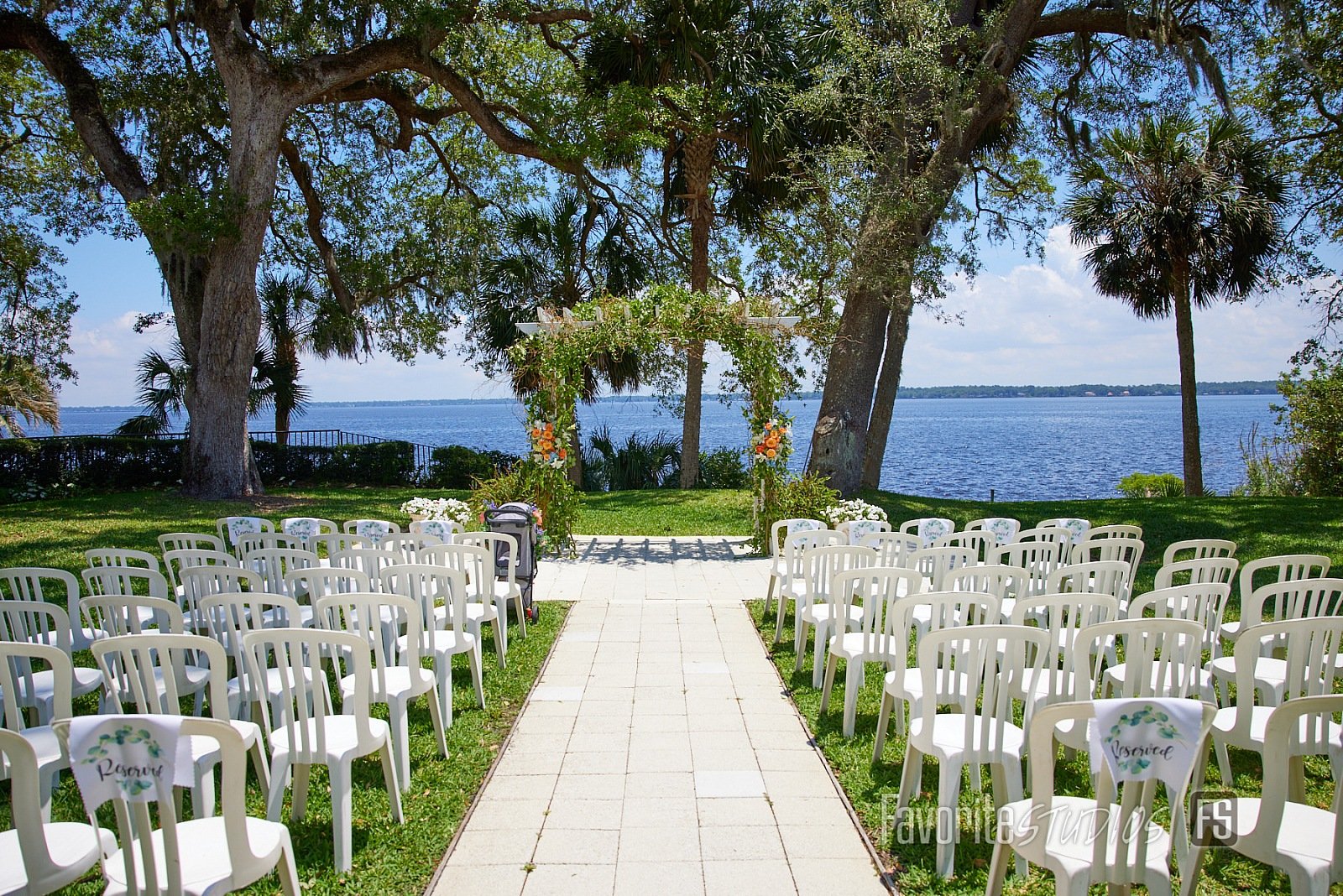 Outdoor wedding ceremony setup with white chairs and floral arch by a lake, surrounded by trees and lush greenery.