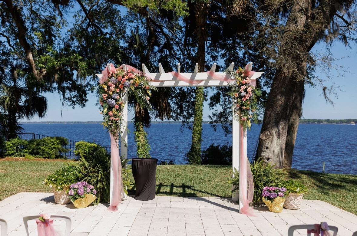Outdoor wedding arch decorated with pink flowers and fabric by a lakeside setting, with trees and water in the background.