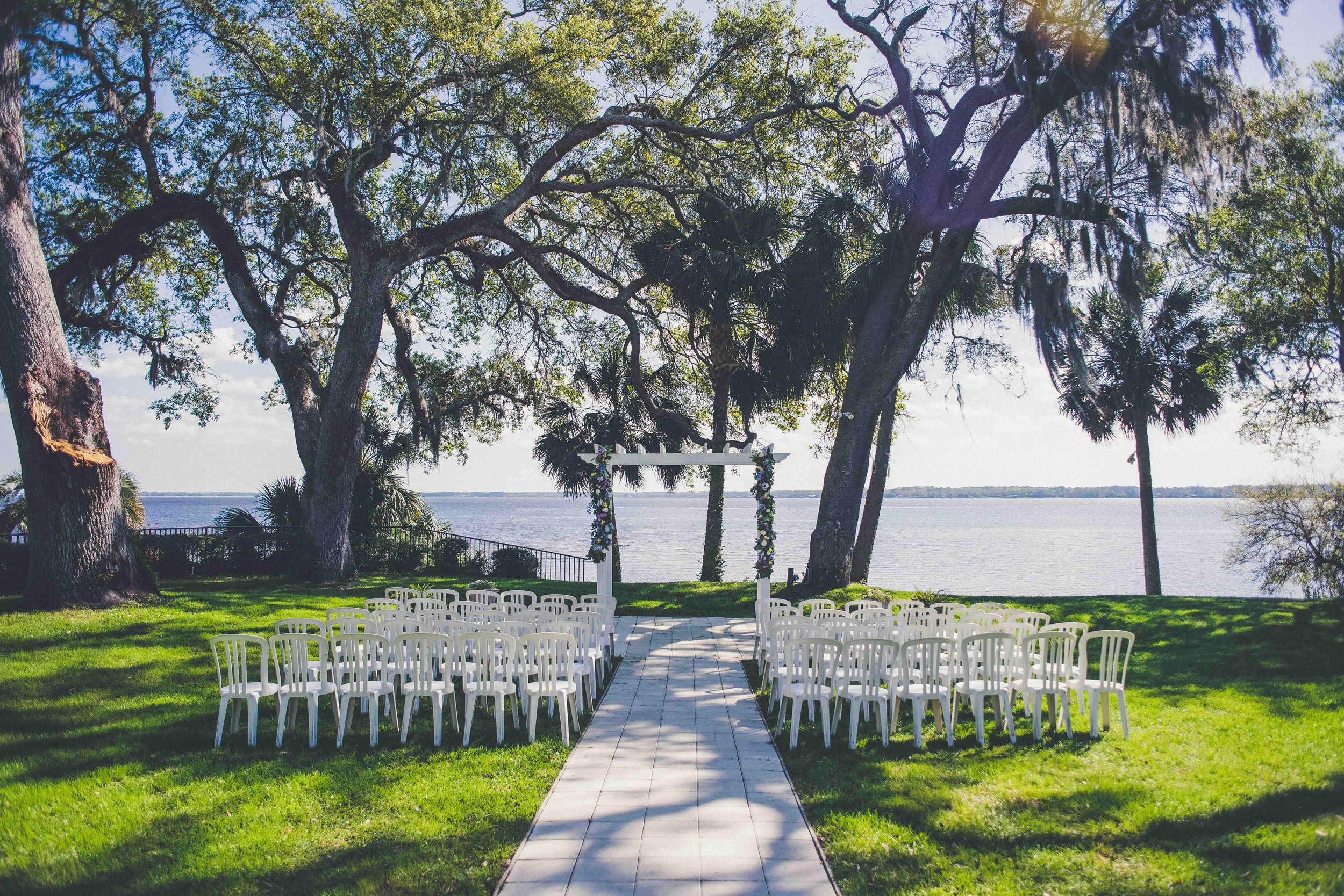 Outdoor wedding setup with white chairs and a decorated arch overlooking a lake, surrounded by large trees and greenery.