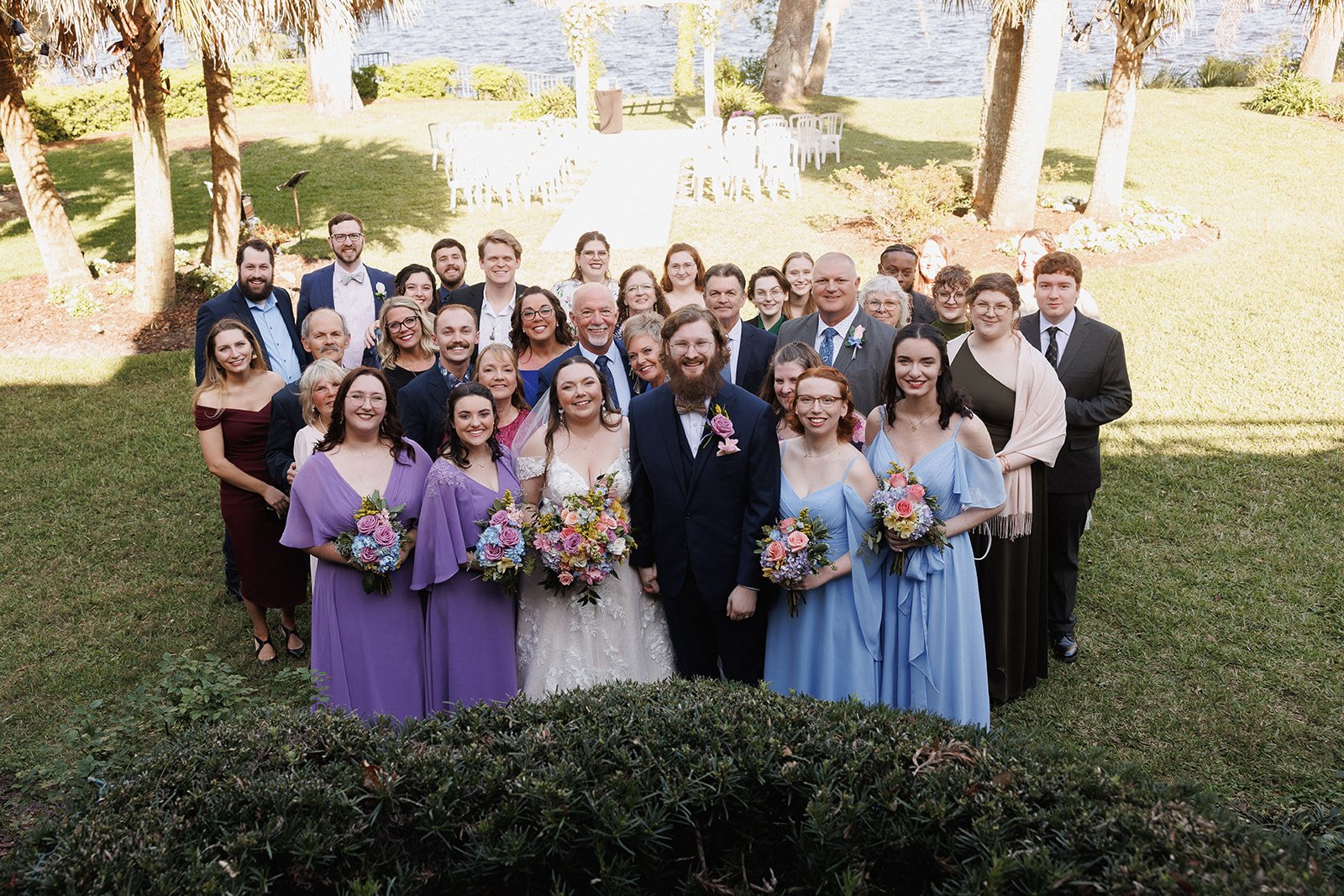 Wedding group photo featuring bride and groom surrounded by bridesmaids in purple and blue dresses, groomsmen, and guests in a garden setting with a water view.