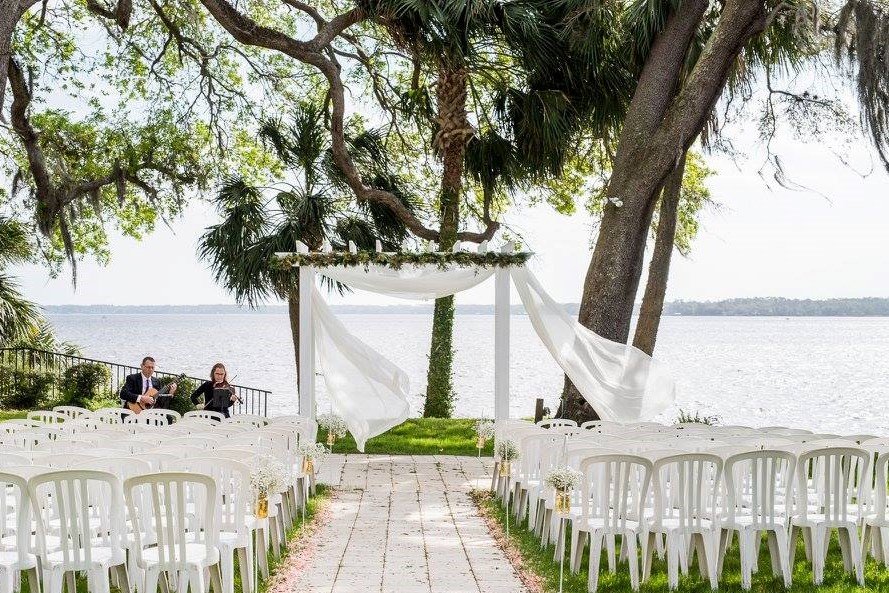 Outdoor wedding ceremony setup by a lake with white chairs, an arch draped with fabric, and musicians playing string instruments.