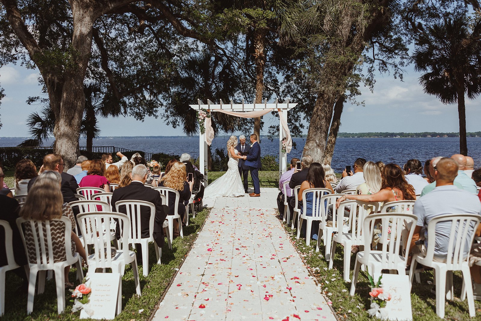 Outdoor lakeside wedding ceremony with chairs, a floral arch, and guests seated on either side of the aisle covered with rose petals.