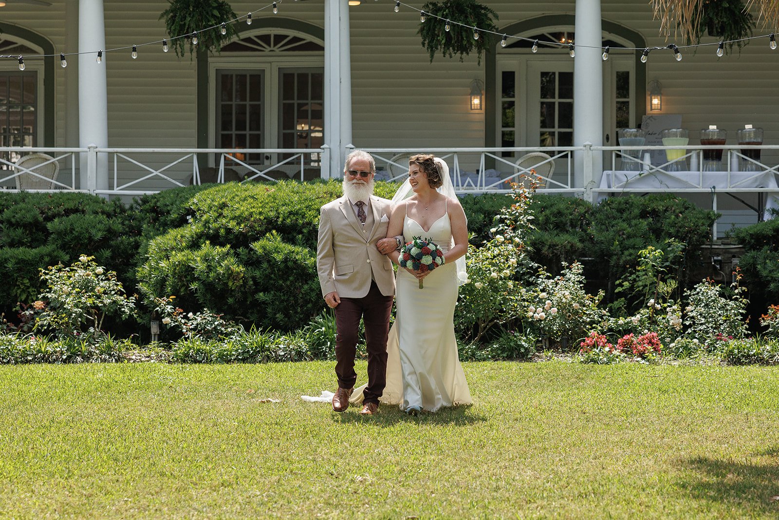 Bride walking down aisle with man in garden setting, holding bouquet, near elegant building with string lights.