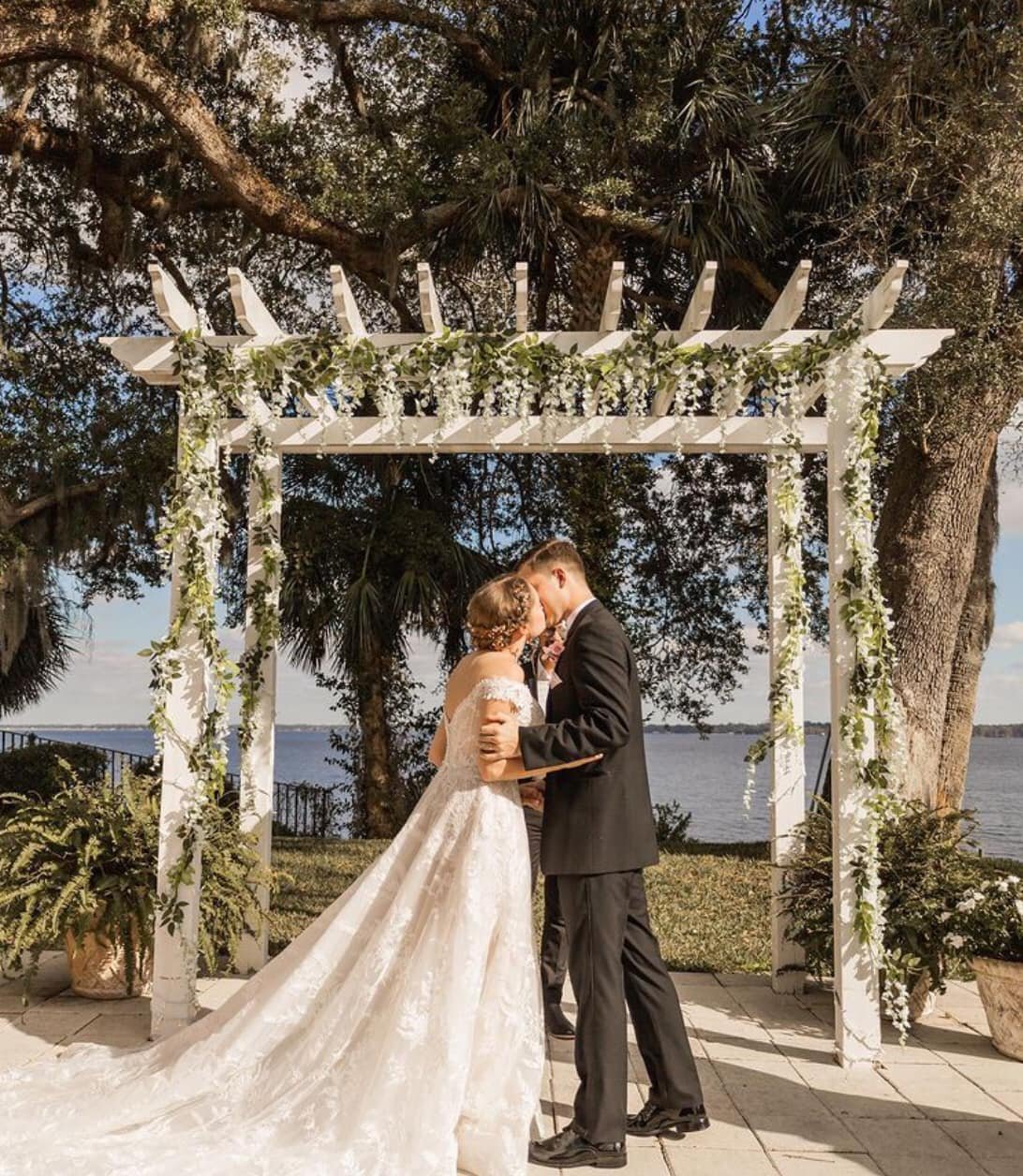 Bride and groom kissing under a white pergola decorated with greenery, near a scenic waterfront, during their outdoor wedding ceremony.
