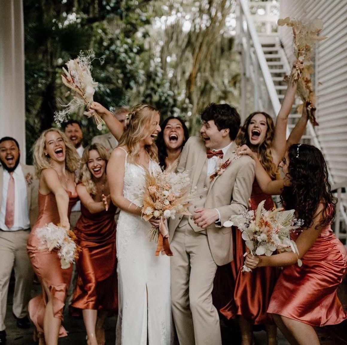 A group celebrating a wedding, featuring a bride in a white dress and a groom in a beige suit surrounded by bridesmaids in terracotta dresses and groomsmen. They're holding floral bouquets and smiling happily.