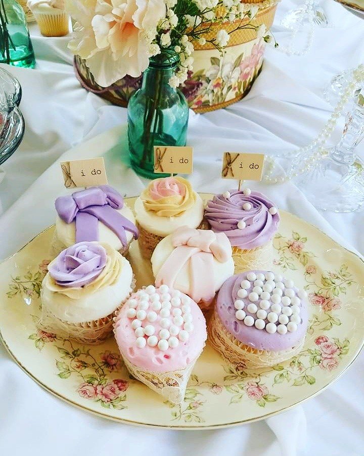 Decorative cupcakes on a floral plate with pastel icing and elegant designs, accompanied by "I do" flags, set as a wedding centerpiece with flowers in the background.