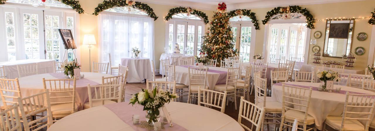 Elegant banquet hall decorated for a holiday event with round tables, white chairs, a Christmas tree, and garlands with lights.