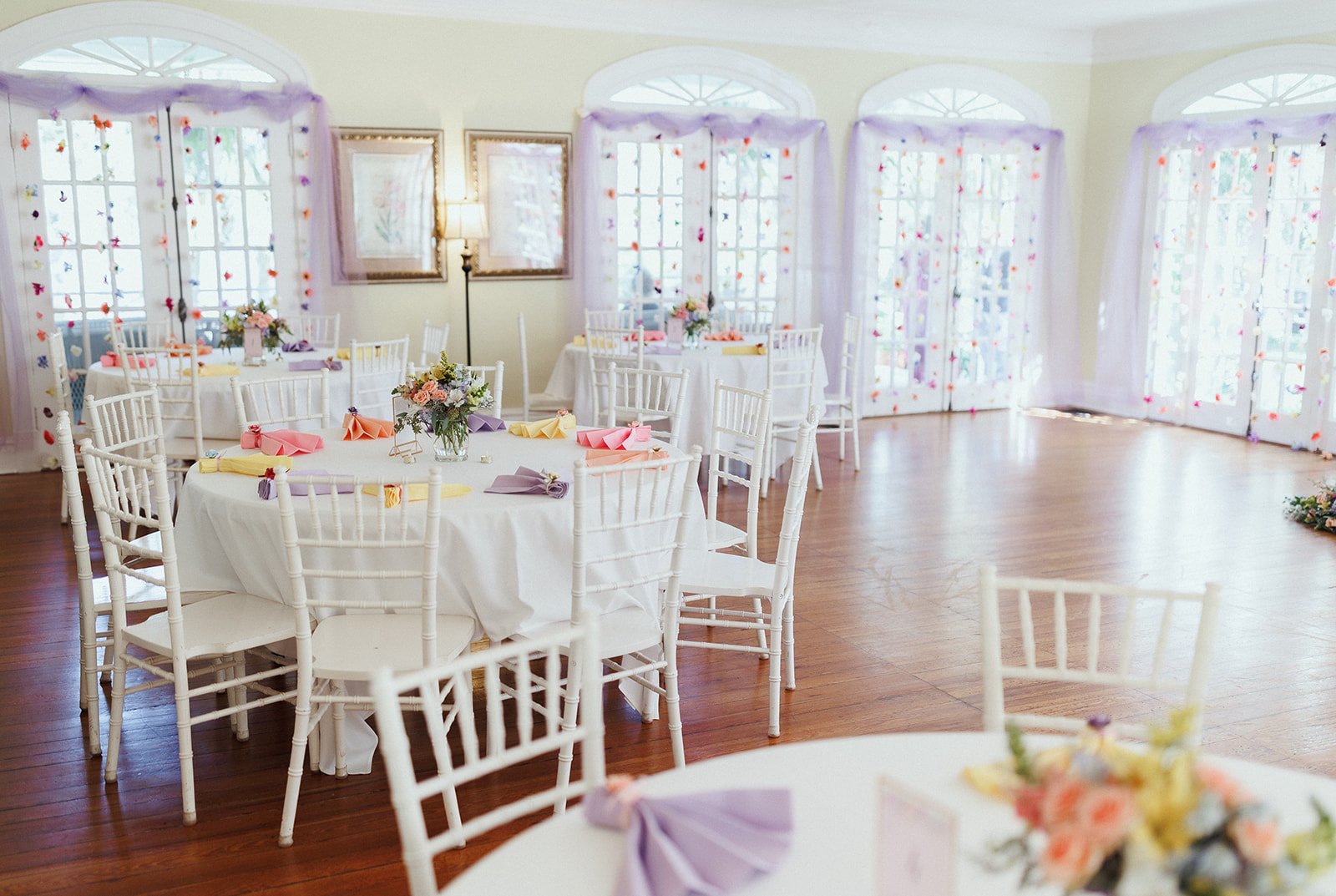 Elegant banquet room with white round tables and chairs, adorned with colorful napkins and floral centerpieces, large windows decorated with lavender curtains and floral accents, hardwood floor.