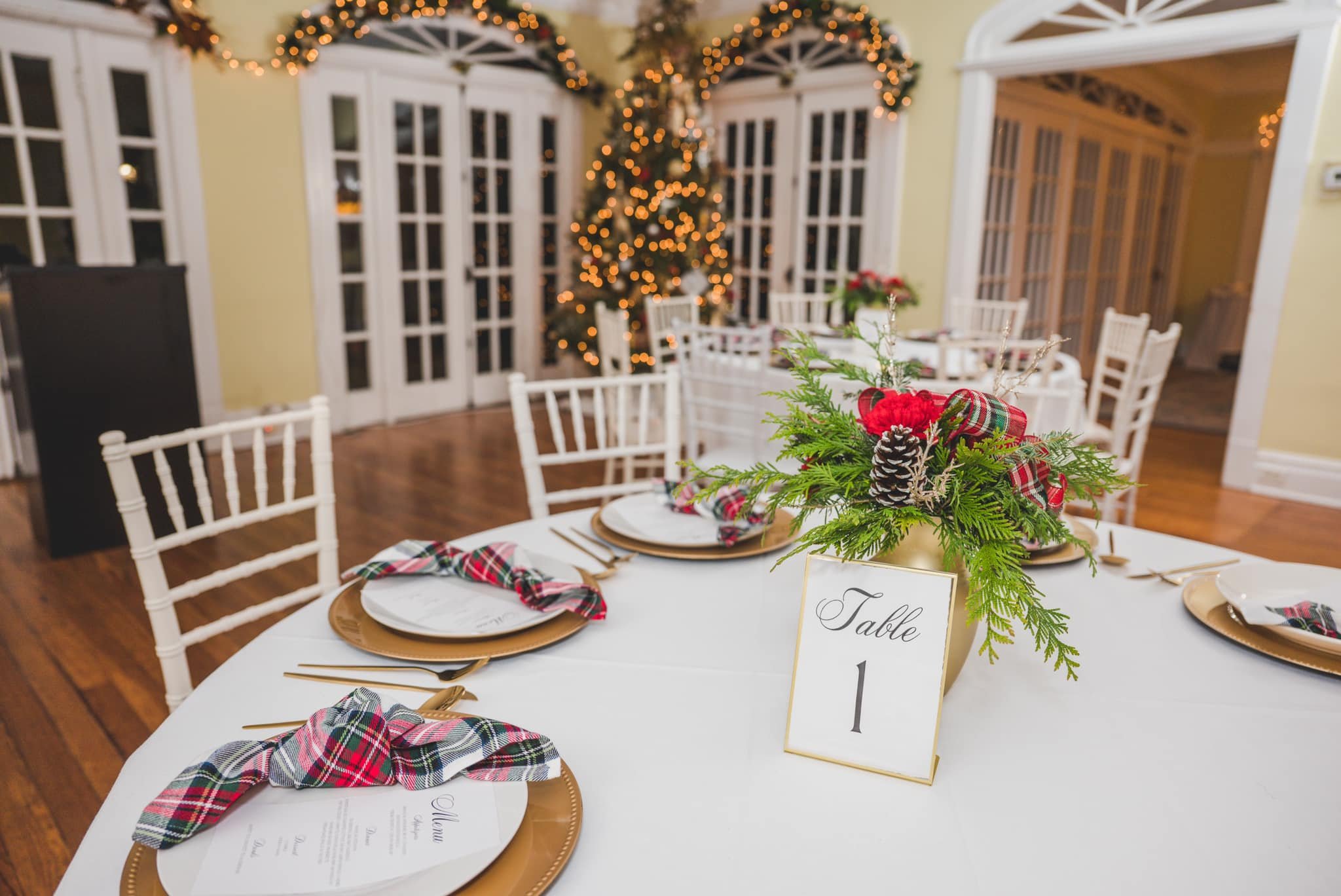 Festive dining table setting with white chairs, decorated with plaid napkins, gold chargers, and a centerpiece of red flowers, greenery, and pinecones. A "Table 1" sign is displayed. Christmas lights and a tree are visible in the background.