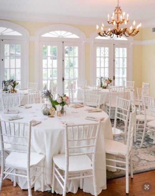 Elegant banquet room with round tables, white tablecloths, white chairs, floral centerpieces, and a chandelier. Large windows and soft wall colors create a bright, inviting atmosphere.
