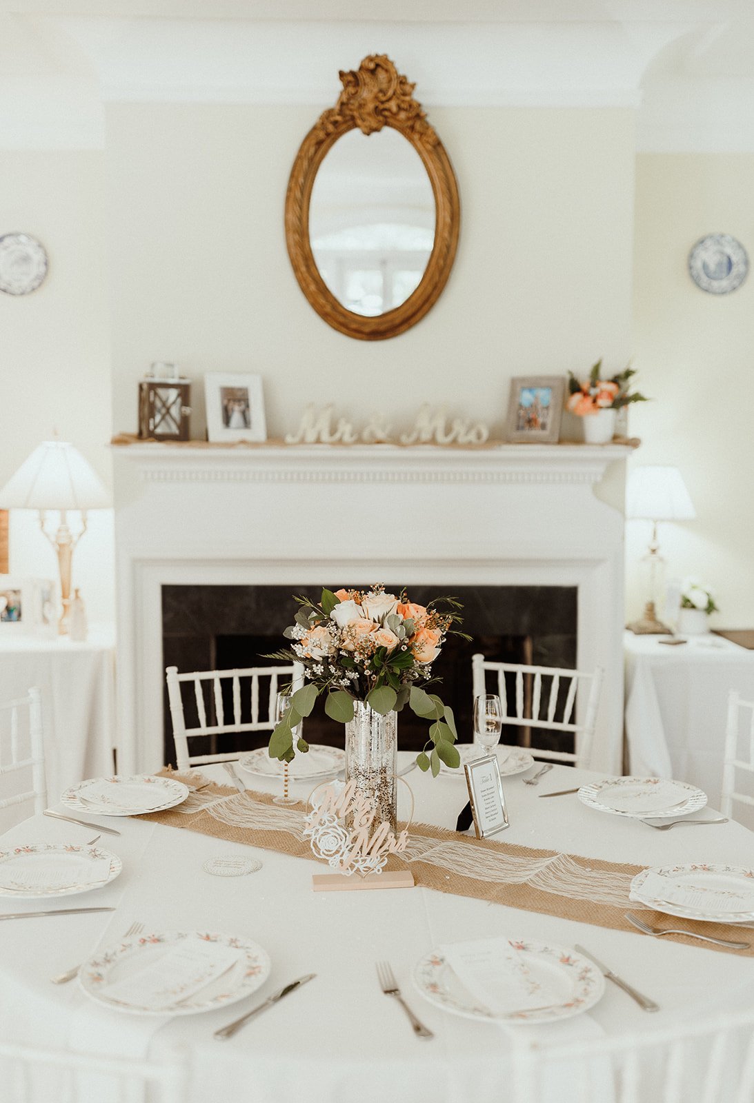 Wedding reception table setup with floral centerpiece, "Mr. & Mrs." sign, and plates on a white tablecloth in a room with a fireplace and ornate mirror.