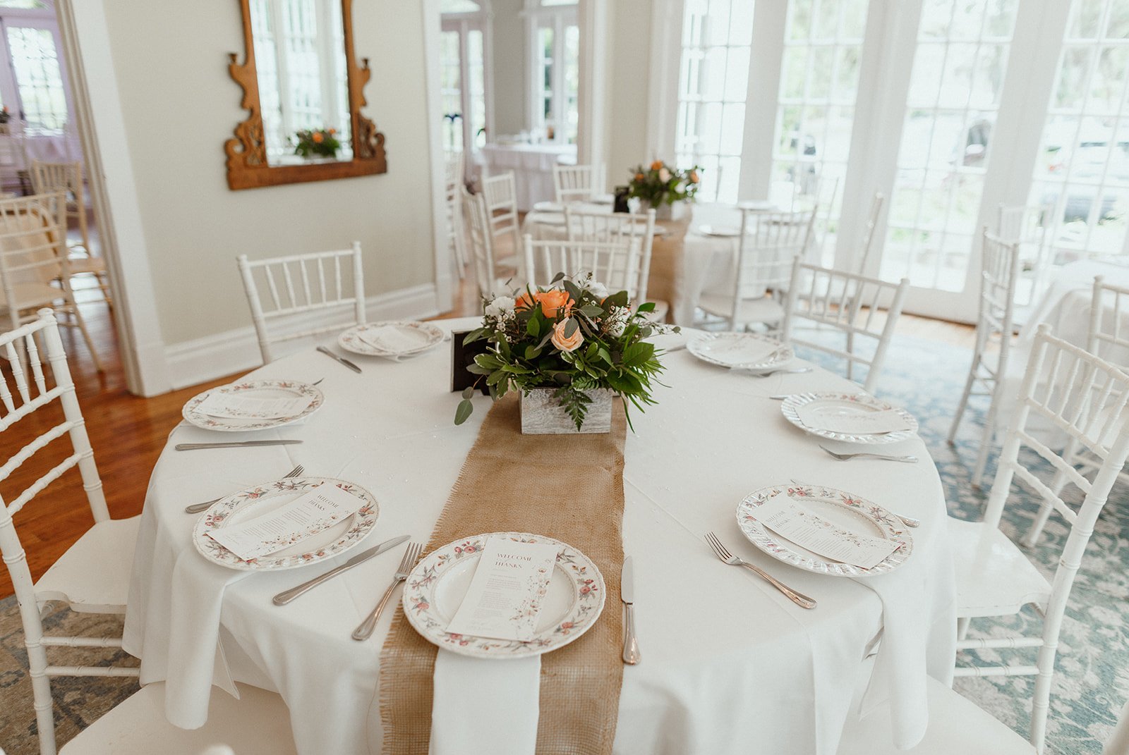 Elegant table setting with floral centerpiece, white tablecloth, burlap runner, floral dinner plates, and white chairs in a bright room with large windows and wooden flooring.