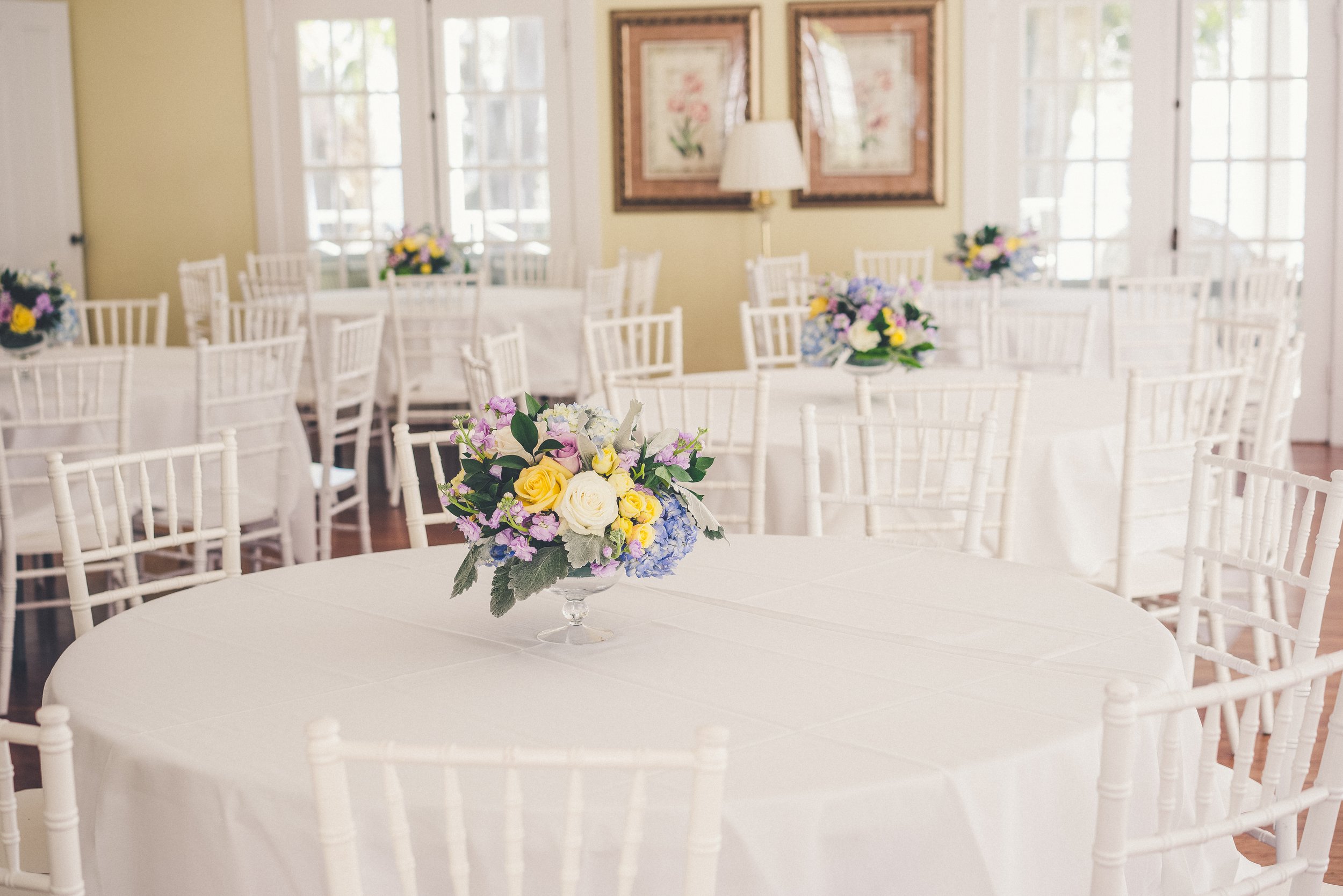 Room with round tables covered in white tablecloths and floral centerpieces, surrounded by white chairs, bright and airy setting with framed art on the walls.