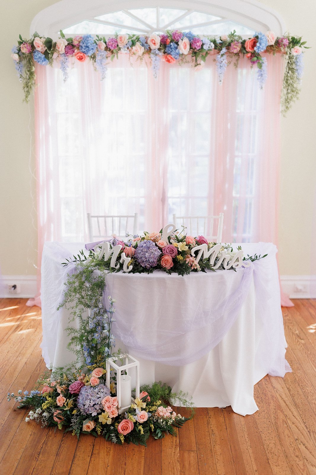 Wedding sweetheart table with "Mr. & Mrs." sign, floral decorations of pink, purple, and blue flowers, under a flower arch, on wooden floor.