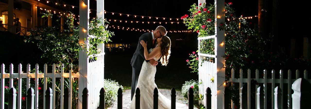 A couple kissing under a lighted archway surrounded by flowers and a white picket fence at night.